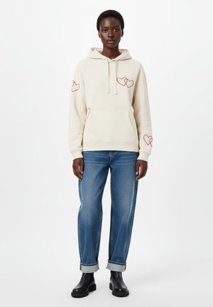 Cream hoodie with red heart outlines on sleeves, front pouch pocket, paired with light blue jeans rolled at cuffs. Black ankle boots.