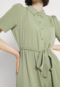 Kobieta w jasnozielonej sukience zapinanej na guziki, z krótkimi rękawami, kołnierzykiem oraz materiałowym paskiem związanym w kokardę w talii.