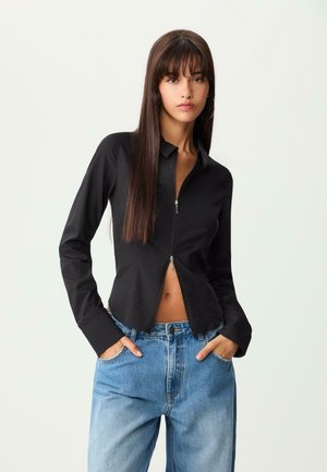 Schwarzes, mit Reißverschluss versehenes Hemd mit Kragen und langen Ärmeln, das eine taillierte Passform hat. Kombiniert mit hochgeschnittenen, hellblauen Jeans.