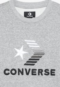 Šedá tkanina s vyšívaným logem Converse, které obsahuje bílou hvězdu, gradientový chevron a černý text "CONVERSE" pod designem.