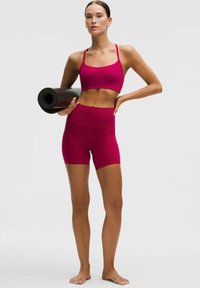 Fukšia športová podprsenka a súprava s vysokým pásom, vyrobená z elastického materiálu. Modelka drží zrolovanú čiernu jogovú podložku.