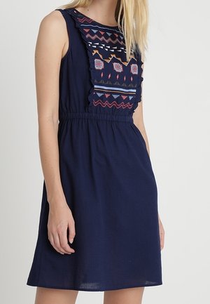 Ärmelloses, marineblaues Kleid mit elastischem Bund und bunter geometrischer Stickerei auf der Brust, getragen von einer stehenden Person mit heller Haut und blonden Haaren.