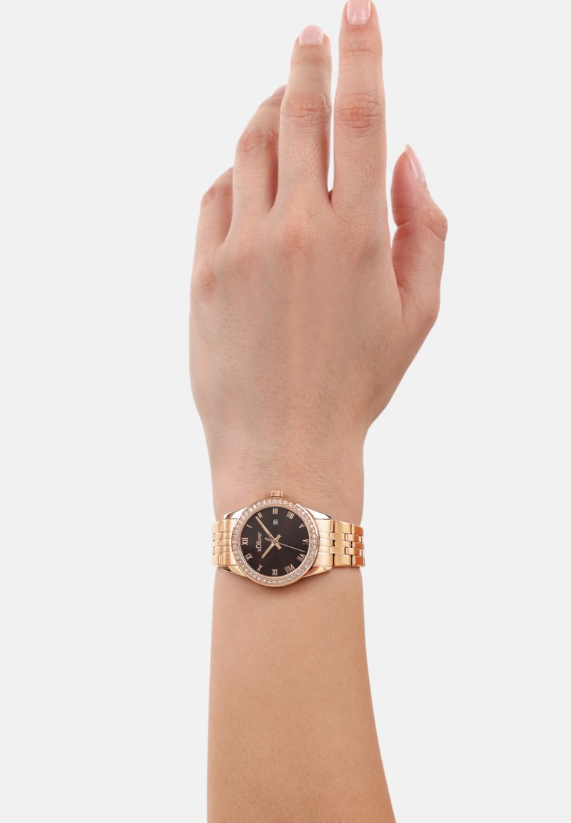 Goldfarbene Uhr mit schwarzem Ziffernblatt, römischen Ziffern und einem mit Kristallen verzierten Rand. Verfügt über ein Metall-Gliederarmband und wird an einer Hand präsentiert.