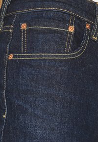 Jean en denim bleu foncé avec des coutures jaunes, des accessoires couleur cuivre et une petite poche avant avec des détails en rivets.