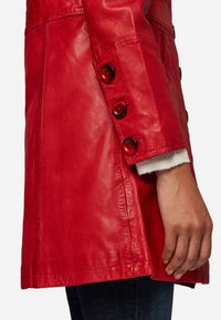 Red leather coat with a slim fit, featuring two large buttons on the cuff and a tailored design. Soft texture with a smooth finish.