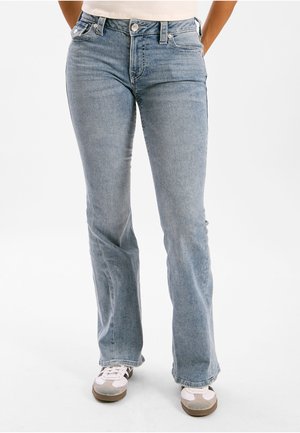 Hellblaue Jeans aus Denim mit leicht ausgestelltem Bein am unteren Saum, getragen mit weißen und grauen Sneakers vor weißem Hintergrund.