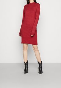 Rode trui jurk met lange mouwen, een ronde hals en een nauwsluitende pasvorm. Gepaarde met zwarte enkellaarsjes met puntige neuzen en een hoekig ontwerp.