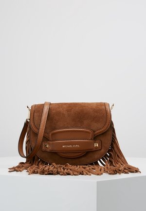 Brun crossbody-taske i ruskind med læderkanter, frynsedetaljer og "Michael Kors"-logo på frontremmen, vist på en hvid piedestal.