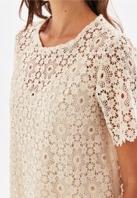 Haut en dentelle avec un motif floral comprenant des motifs circulaires, un décolleté en bordure festonnée et des manches courtes de couleur beige clair.