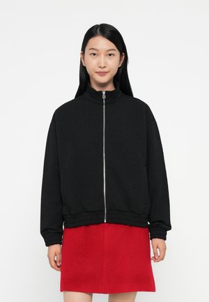 Black zip-up jacket with a textured pattern, mock collar, and elastic cuffs. Paired with a red mid-length skirt. Simple, modern design.
