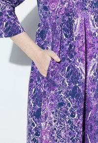 Robe violette à motifs avec un design marbré. Les longues manches et les poches latérales améliorent la fonctionnalité. La texture du tissu lisse est visible.