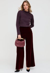 Weite Hosen aus burgunderrotem Samt, verkürzter lila Pullover und eine gewebte burgunderrote Handtasche mit goldenem Griff. Schwarze spitze High Heels.