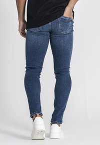 Jeans ajustados de mezclilla azul oscuro con una textura suave, que presentan una cintura de altura media y bolsillos traseros. Combinados con zapatillas blancas.