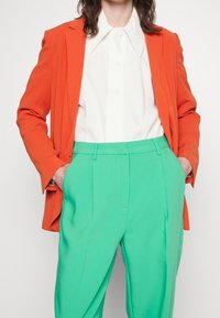 Vrouw in een wit shirt, feloranje blazer en hooglopende groene broek met handen in de zakken tegen een effen achtergrond.