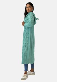 Langer, mit Kapuze versehener Cardigan in helltürkis, mit Zopfmusterstruktur und geripptem Saum. Kombiniert mit schmal geschnittenen Jeans und lässigen Sneakers.