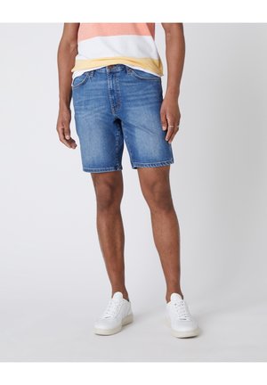 Mann trägt blaue Jeans-Shorts, weiße Sneakers und ein pastellfarben gestreiftes T-Shirt und steht vor einem einfarbigen Hintergrund.