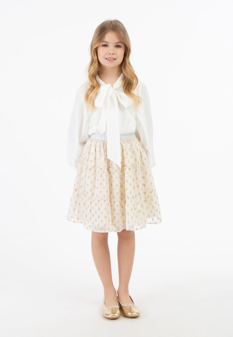 Witte blouse met een strik, gelaagde crèmekleurige rok met gouden patronen, zilveren tailleband en glanzende gouden ballerina's.