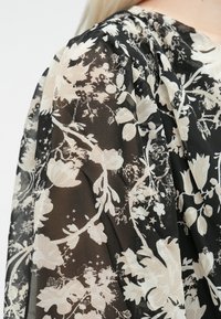 Tessuto nero trasparente con un motivo floreale avorio caratterizzato da texture variabili e un design fluido. I dettagli decorativi includono sottili stelle.