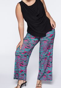 Svart ärmlös topp med draperad halsringning, tillsammans med vida byxor med ett mönster av tigerrand i teal och rosa. Svart sandaler med remmar.