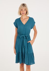 Teal, korte mouwen jurk van gekreukt materiaal. Heeft een diepe V-hals, een taille met strik, en een uitlopende, gelaagde rok. Inclusief zakken.