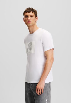 White, short-sleeve t-shirt made of cotton, featuring a textured graphic design on the front. Shown with gray denim pants.