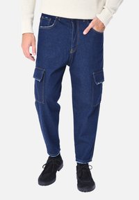Pantaloni cargo in denim blu con ampie tasche laterali, cuciture a contrasto e vestibilità rilassata. Indossati con calzature nere. La texture è liscia e resistente.