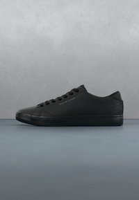 Baskets basses noires avec des lacets et le logo Tommy Hilfiger sur le côté, exposées sur une surface lisse sur fond gris.