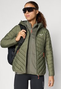 Olīvzaļš pūkains jaka ar čevronu quilted rakstu, oranžiem rāvējslēdzējiem, valkāta uz teksturētas pelēkas blūzes. Melna mugursoma un saulesbrilles.