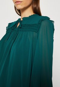 Femme portant un chemisier vert foncé transparent avec empiècement plissé, col volanté et fermeture à nouer au niveau du cou sur un fond uni.