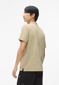 Béžová polokošeľa s krátkym rukávom vyrobená z mäkkého bavlneného materiálu, s golierom, bočnými rozparkom a vo voľnom strihu, nosená s čiernymi cargo nohavicami.