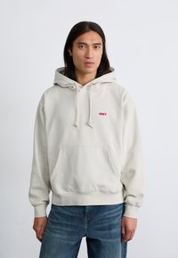 Light beige hoodie made of cotton, featuring a front pocket, drawstrings, and a small red "OBEY" logo on the chest. Styled with blue jeans.