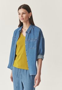Denim košulja na dugih rukava svijetloplave boje s podvrnutim rukavima, u kombinaciji s vibrantnom žutom majicom s V-izrezom i ležernim trapericama.