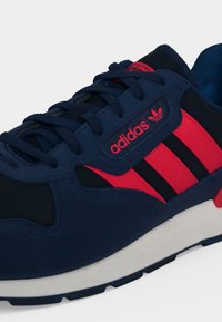 Σκούρο μπλε και κόκκινο αθλητικό παπούτσι Adidas με μαύρες ρίγες, κορδόνια και λευκή σόλα, με το λογότυπο Adidas στην πλαϊνή όψη.