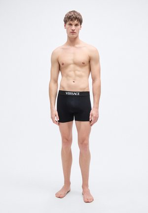 Μαύρα ανδρικά εσώρουχα μποξέρ με ελαστική μέση με λογότυπο. Το ύφασμα φαίνεται λείο και εφαρμοστό. Το μοντέλο στέκεται ξυπόλυτο με ουδέτερη έκφραση.