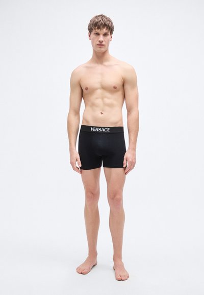 Caleçon boxer noir avec une ceinture élastique de marque. Le tissu semble lisse et ajusté. Le mannequin est pieds nus avec une expression neutre.