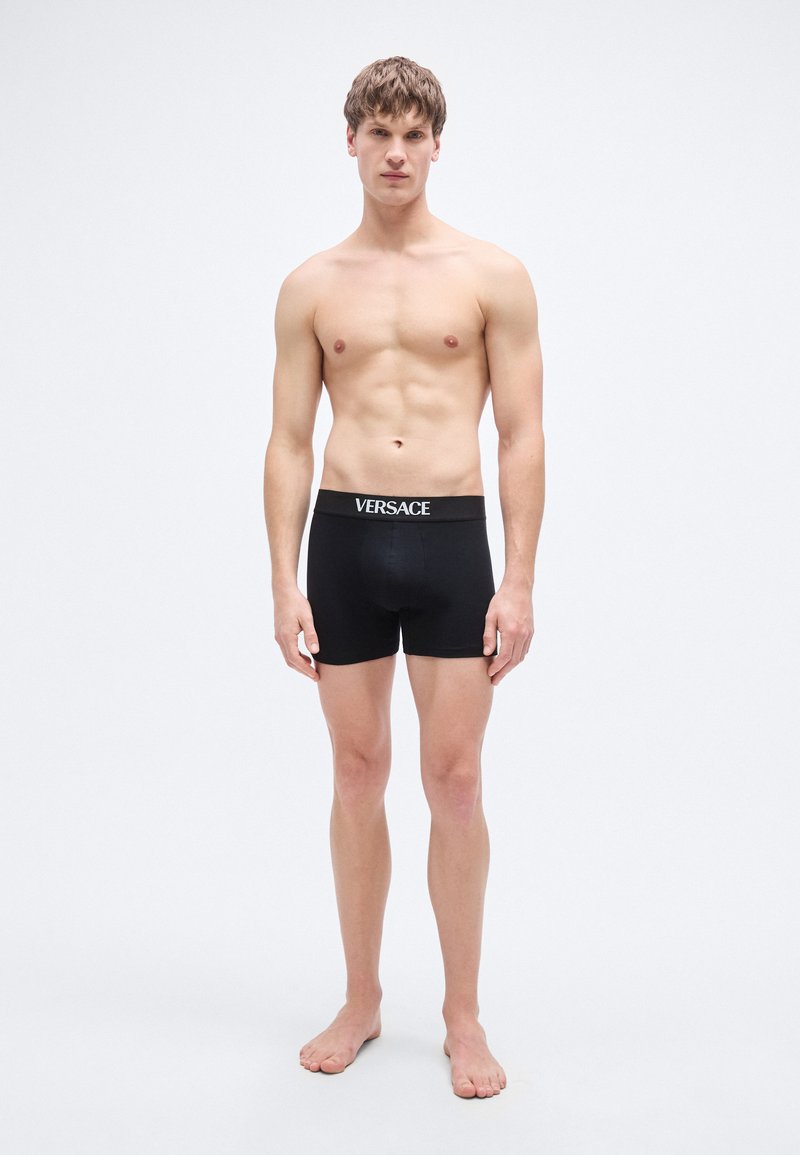 Caleçon boxer noir avec une ceinture élastique de marque. Le tissu semble lisse et ajusté. Le mannequin est pieds nus avec une expression neutre.