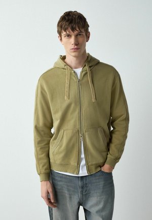 Mladý muž v olivovo zelenej mikine na zips s tiahadlami a modrých džínsoch stojaci pred jednoduchým svetlým pozadím.