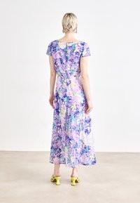 Kwiatowa sukienka maxi w odcieniach fioletu, niebieskiego i żółtego; krótkie rękawy, rozcięcie z tyłu, zwiewny materiał, zestawiona z żółtymi sandałami na obcasie.
