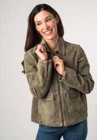 Giacca in suede verde oliva con colletto, chiusura frontale con zip e due tasche applicate. Texture morbida con dettagli cuciti.