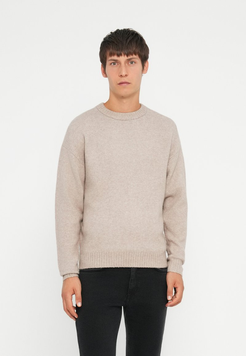 Light beige knit sweater with a classic crew neckline, ribbed cuffs and hem, and a soft, textured finish. Worn with black jeans.