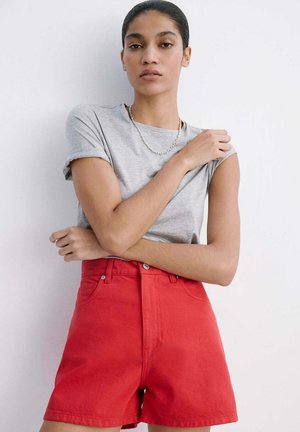 Mujer con camiseta gris y pantalones cortos rojos de talle alto, de pie con los brazos cruzados contra un fondo blanco.