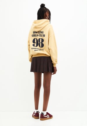 Person mit dem Rücken zur Kamera, trägt einen gelben Hoodie mit dem Text "Bowling Girls Club 98 Nashville", karierten Rock, weiße Socken und bordeauxrote Sneaker.