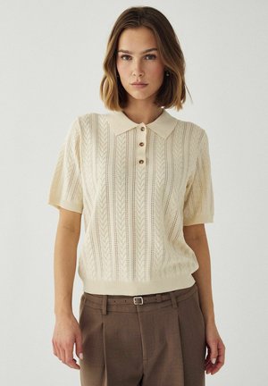 Kvinne iført kremfarget kortermet strikket polo med knapper og brune høytlivsbukser med belte, stående mot en enkel lys bakgrunn.