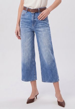 Vrouw draagt lichtblauwe wijde cropped jeans met een bruine riem en bruine pumps met een spitse neus, staand tegen een effen achtergrond.