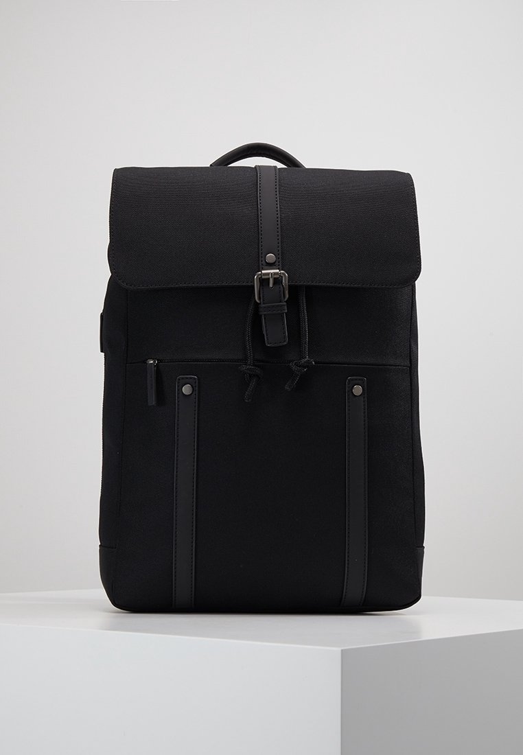 Sac à dos en tissu noir avec un rabat sécurisé par une boucle, poche avant zippée, fermeture à cordon et poignée supérieure sur un socle blanc.