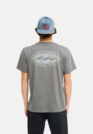 Persoon draagt een grijs Billabong T-shirt met golflogo en tekst "Since 1973" op de achterkant, gecombineerd met een blauwe denim pet en zwarte broek.