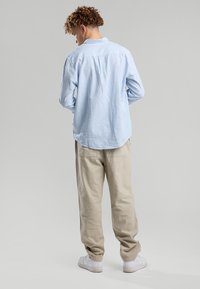 Young man with curly hair wears a light blue striped shirt, beige pants, and white sneakers, standing with hands in pockets facing away.