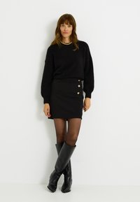 Suéter negro de felpa con mangas largas, combinado con una minifalda negra con acentos de botones dorados. Botas negras hasta la rodilla completan el look.