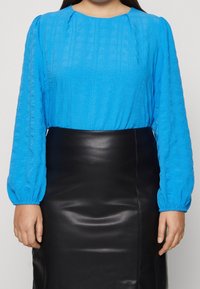 Blaue Texturoberteil mit langen Puffärmeln, kombiniert mit einem schwarzen Leder-Bleistiftrock. Das Oberteil hat ein geripptes Muster und einen runden Ausschnitt.