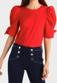 Kvinde iført en lys rød top med puffærmer og bundet manchetter, samt højtaljede mørkeblå jeans med seks sølvknapper.